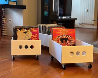 以下が含まれることがあります： 黒い車輪が付いた2つの白い木製レコード収納カート。各カートの前面には3つの円形の切り抜きがあります。カートはビニールレコードで満たされています。