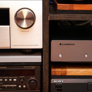May include: A close-up of a home entertainment system with a silver receiver, a black and silver VCR, and a silver amplifier with the text "CAMBRIDGE" on it. The system is on a wooden shelf.