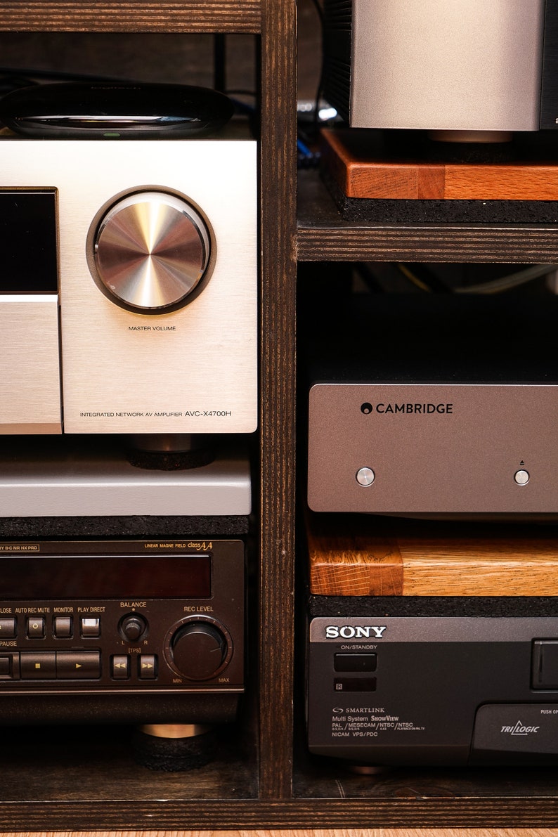 May include: A close-up of a home entertainment system with a silver receiver, a black and silver VCR, and a silver amplifier with the text "CAMBRIDGE" on it. The system is on a wooden shelf.
