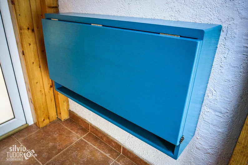 May include: A blue wall-mounted folding table with a shelf underneath. The table is closed and has a simple design.