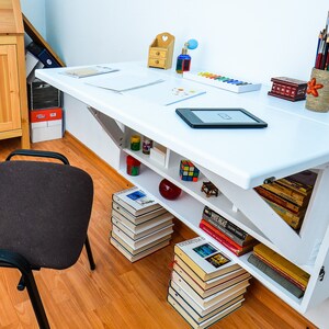 May include: White wall-mounted folding desk with a white tabletop and shelves. The desk has a small shelf with books and a larger shelf with a stack of books. There is a small wooden box, a bottle of paint, and a set of paintbrushes on the desk.