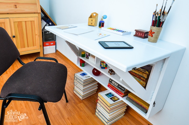 May include: White wall-mounted folding desk with a white tabletop and shelves. The desk has a small shelf with books and a larger shelf with a stack of books. There is a small wooden box, a bottle of paint, and a set of paintbrushes on the desk.