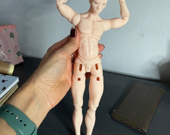 Puede incluir: Una muñeca articulada de color carne con un cuerpo de tipo masculino. La muñeca está en pose con los brazos levantados por encima de la cabeza. La muñeca tiene una apariencia realista con músculos esculpidos y una cara detallada.