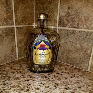 May include: A clear glass bottle of Crown Royal Fine De Luxe Blended Canadian Whisky. The bottle has a gold label with a purple crown and the words "Crown Royal" in gold lettering.