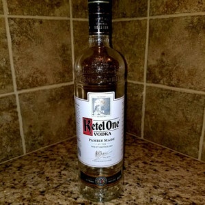 May include: A clear glass bottle of Ketel One Vodka. The bottle has a black label with white text that reads "Ketel One Vodka Family Made at the Nolet Distillery."