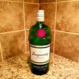 May include: A green glass bottle of Tanqueray London Dry Gin, with a white label and silver cap. The label reads "Tanqueray Imported London Dry Gin". The bottle has a red wax seal and is displayed on a speckled countertop.