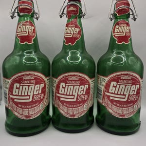 May include: Three green glass bottles of "Ginger Brew" with red and white labels. Each bottle has a swing top closure and a white stopper. The labels read "Sparkling Ginger Brew" and "NET 25.36 FL OZ (750 ml)".