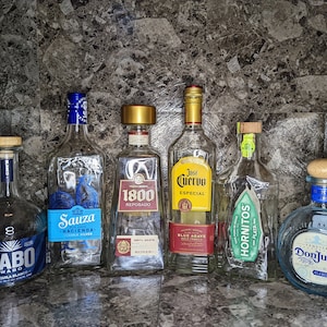 May include: A collection of seven clear glass bottles of tequila, each with a different brand label. The bottles are arranged in a row on a textured surface. The brands include Cabo Wabo, 1800 Reposado, Sauza Hacienda, Jose Cuervo Especial, Hornitos Plata, and Don Julio Blanco.