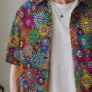 May include: A short-sleeved button-down shirt featuring a vibrant, all-over print of multi-colored stylized flowers with smiling faces. The shirt is worn open over a plain white t-shirt, paired with light tan trousers and a simple silver necklace.