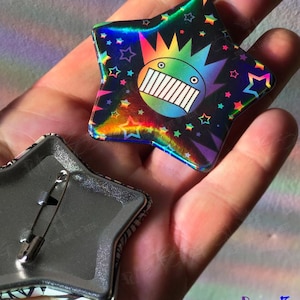 May include: Star-shaped button with a holographic finish, featuring a cartoon face with a rainbow design and a spiky crown. The button has a pin back. The text "Put a Foot on it" is visible in the bottom right corner.