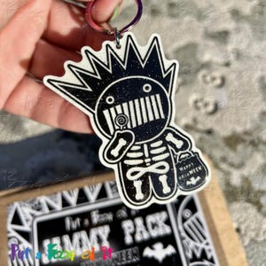 May include: A black and white acrylic keychain featuring a stylized character with spiky hair, a skeleton ribcage design, and a lollipop. The character holds a small bucket labeled Happy Halloween. The surface has a subtle glitter finish.