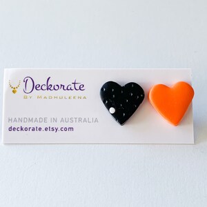 May include: A pair of heart-shaped earrings. One earring is black with white dots and a white accent, the other is bright orange. The earrings are displayed on a white card with the text "Deckorate by Madhuleena" and "Handmade in Australia".