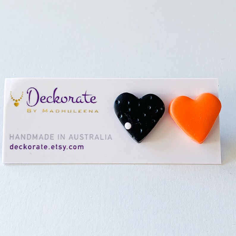 May include: A pair of heart-shaped earrings. One earring is black with white dots and a white accent, the other is bright orange. The earrings are displayed on a white card with the text "Deckorate by Madhuleena" and "Handmade in Australia".