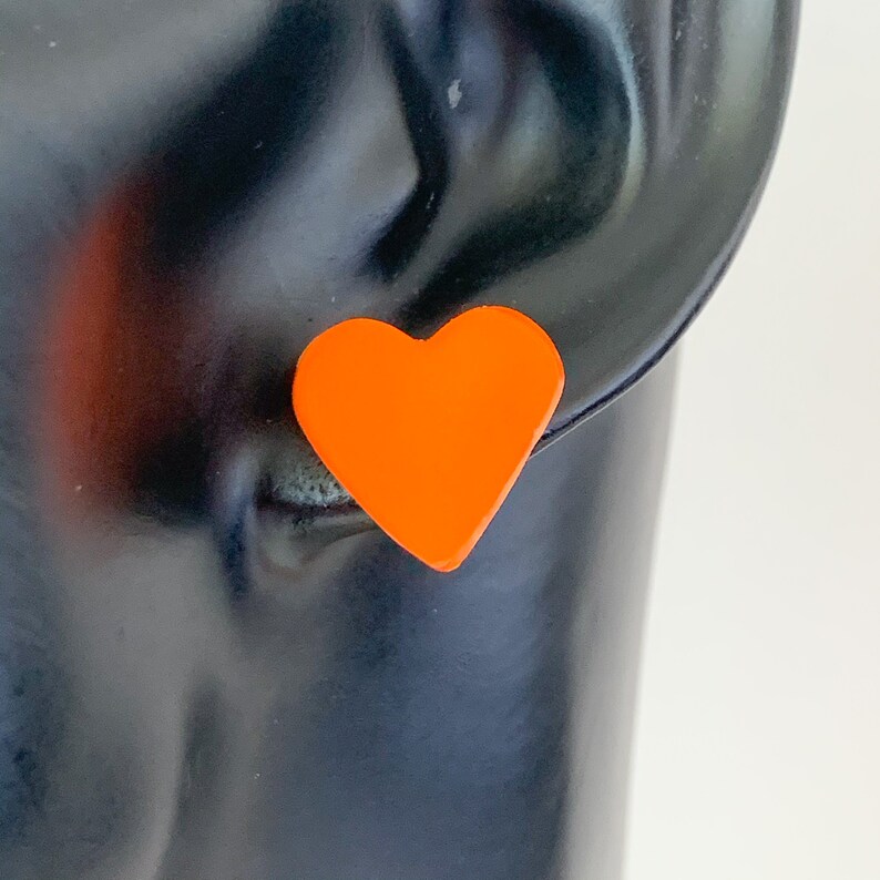 May include: Bright orange heart-shaped stud earrings. The earrings are a solid, vibrant orange color and are set against a black background. These earrings are a simple yet bold accessory.