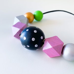 Pu&ograve; includere: Una collana colorata con perline di legno dipinte in varie forme e colori. La collana include una perlina blu navy a pois bianchi, perline geometriche lavanda e argento, e perline sferiche dorate e verdi. Le perline sono infilate su un cordoncino nero.