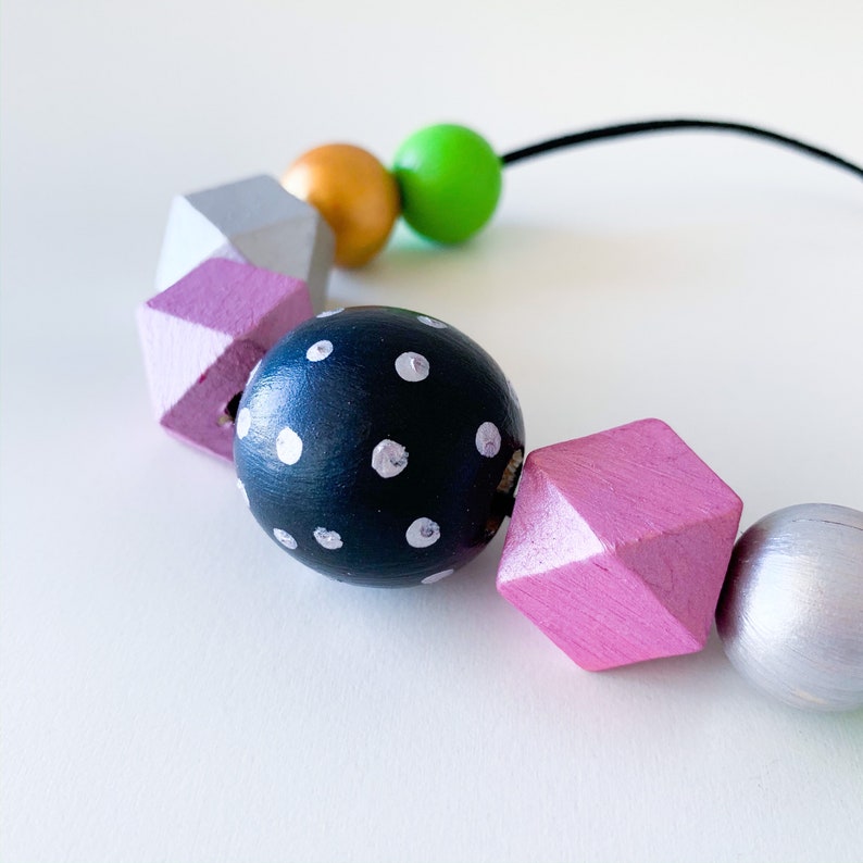 Pu&ograve; includere: Una collana colorata con perline di legno dipinte in varie forme e colori. La collana include una perlina blu navy a pois bianchi, perline geometriche lavanda e argento, e perline sferiche dorate e verdi. Le perline sono infilate su un cordoncino nero.