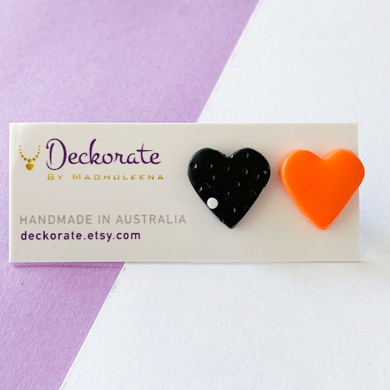 May include: A pair of heart-shaped earrings. One earring is black with white dots and a single white circle. The other earring is bright orange. The earrings are displayed on a white card with the text "Deckorate by Madhuleena" and "Handmade in Australia".