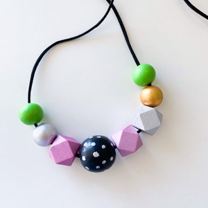 Pu&ograve; includere: Una collana colorata con un cordino nero e perline di legno dipinte. Le perline sono in vari colori: verde, argento, oro, rosa e una perlina nera a pois bianchi. La collana ha un design geometrico moderno.