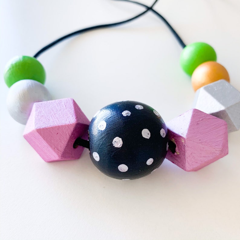 Pu&ograve; includere: Una colorata collana di perline di legno con una variet&agrave; di forme e colori. La collana include una grande perla nera a pois, perline rosa geometriche e perline rotonde in verde, arancione e argento. Infilata su un cordino nero.