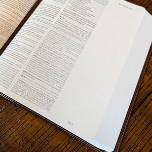 Puede incluir: Biblia abierta con cubierta de cuero marr&oacute;n, mostrando texto en las p&aacute;ginas. Los bordes de las p&aacute;ginas son blancos y el libro est&aacute; abierto sobre una superficie de madera. El n&uacute;mero de p&aacute;gina 1251 es visible.