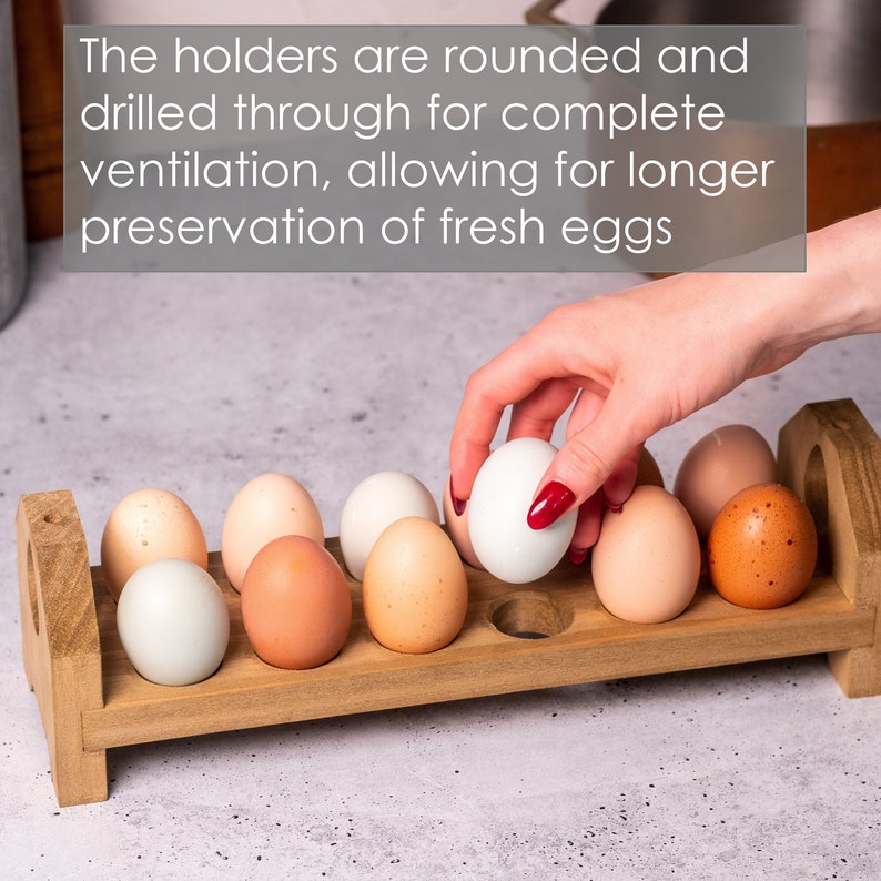 May include: A wooden egg holder with rounded holes to allow for ventilation. The holder is filled with a variety of brown, white, and blue eggs. A hand is placing a white egg into the holder.