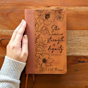 May include: A brown leather-bound book with floral engravings and the text "She is clothed with strength & dignity" and "Proverbs 31:25". The book has a ribbon bookmark and is personalized with the name "Kate Thompson".