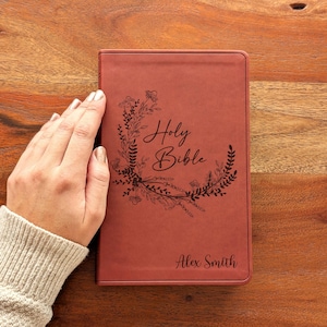 May include: A reddish-brown leather-bound Bible with the words "Holy Bible" and the name "Alex Smith" embossed on the cover. The text is surrounded by a floral wreath design.