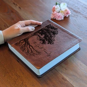 Puede incluir: Un libro encuadernado en cuero marr&oacute;n con un dise&ntilde;o de &aacute;rbol y el texto "The Smith Family" grabado en la portada. El libro tiene p&aacute;ginas con bordes blancos y un borde cosido. Un peque&ntilde;o ramo de flores rosas y blancas descansa cerca.