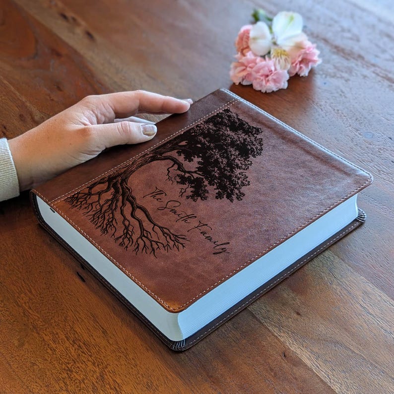 Puede incluir: Un libro encuadernado en cuero marr&oacute;n con un dise&ntilde;o de &aacute;rbol y el texto "The Smith Family" grabado en la portada. El libro tiene p&aacute;ginas con bordes blancos y un borde cosido. Un peque&ntilde;o ramo de flores rosas y blancas descansa cerca.