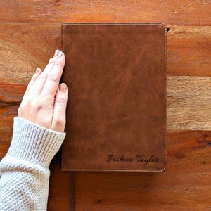 May include: A brown leather journal with the name "Joshua Taylor" embossed on the lower right corner. The journal has a ribbon bookmark and is set against a wooden background.