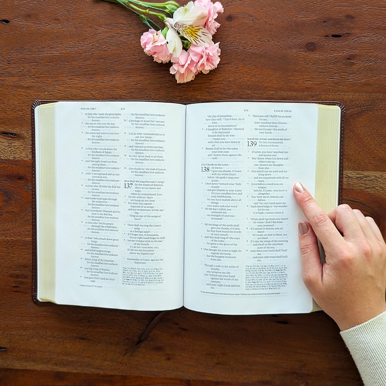 May include: An open Bible with a brown leather cover, text-filled pages, and a finger indicating a passage. Pink and white flowers are in the upper left corner. The book rests on a wooden surface.