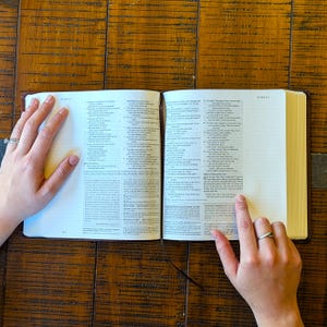 Puede incluir: Biblia abierta con texto visible en las p&aacute;ginas. El libro tiene una cubierta de cuero marr&oacute;n y est&aacute; abierto sobre una superficie de madera. Una mano con un anillo se&ntilde;ala una l&iacute;nea de texto.