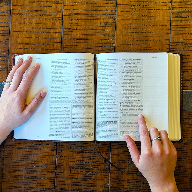 Puede incluir: Biblia abierta con texto visible en las p&aacute;ginas. El libro tiene una cubierta de cuero marr&oacute;n y est&aacute; abierto sobre una superficie de madera. Una mano con un anillo se&ntilde;ala una l&iacute;nea de texto.
