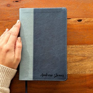 Puede incluir: Un diario de dos tonos, gris y azul marino, con el nombre "Andrew Jones" grabado en la portada. El diario está sostenido por una mano, y la portada tiene un borde cosido.