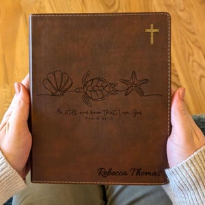 May include: Brown leather Bible cover with a gold cross and a line drawing of a seashell, turtle, and starfish. The cover has the text "Be still and know that I am God" and the name "Rebecca Thomas".