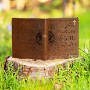 May include: A brown leather Bible cover, open to display the interior. The cover features a sunflower design and the text "God is within her. SHE will not fall." Additional text includes a Bible verse and a name.