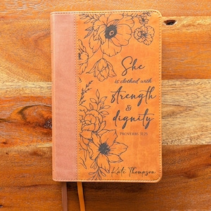 Könnte beinhalten: Ein ledergebundenes Buch in Hellbraun mit floralen Illustrationen und dem Text "She is clothed with strength & dignity" und "Proverbs 31:25". Das Buch hat einen pfirsichfarbenen Buchrücken und ein passendes Lesezeichen. Der Name "Kate Thompson" steht unten.