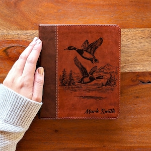 May include: A brown leather journal with a dark brown spine. The cover features an engraved illustration of two ducks in flight over a mountain landscape. The name "Mark Smith" is printed at the bottom.