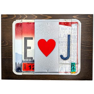 May include: A framed wall art piece featuring license plates, a red heart, and a blue letter "J". The artwork includes the letter "E" and the number "12" on the license plates. The frame is made of dark brown wood.
