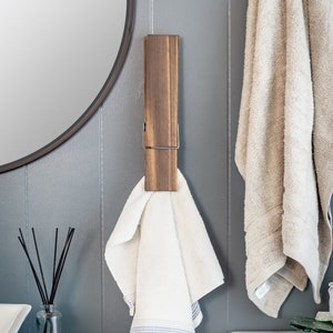 Puede incluir: Dos toalleros de madera con clips met&aacute;licos, montados en una pared gris. Un toallero tiene una toalla blanca colgada, el otro tiene una toalla beige colgada.