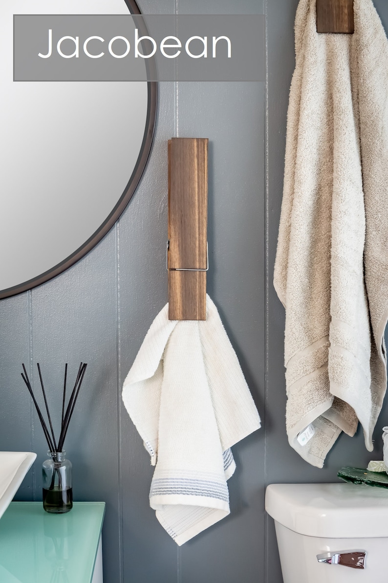 Puede incluir: Dos toalleros de madera con clips met&aacute;licos, montados en una pared gris. Un toallero tiene una toalla blanca colgada, el otro tiene una toalla beige colgada.