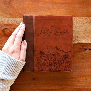 Pode incluir: Uma Bíblia em couro castanho com as palavras "THE Holy Bible" em escrita cursiva. A capa apresenta um desenho floral e o nome "Taylor Dunshee". A Bíblia é segurada por uma pessoa que usa um suéter cinza.