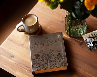 May include: A leather-bound Bible with the title "The Holy Bible" and the name "Taylor Smith" is displayed on a wooden table. The cover features an engraved floral design and a cross. A cup of coffee and a vase of flowers are also on the table.