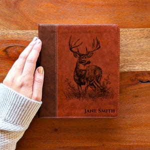 May include: A brown leather journal with a deer illustration and the name "JANE SMITH" printed on the cover. The journal has a dark brown spine and is held by a person wearing a gray sweater.