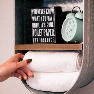 May include: A galvanised metal wall shelf with two shelves. The top shelf has a small box with the text "You never know what you have until it's gone. Toilet paper, for instance." and a small glass jar. The bottom shelf has a white alarm clock with a green face and a stack of white towels.