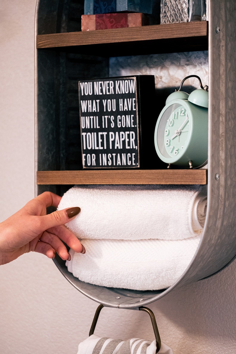 May include: A galvanised metal wall shelf with two shelves. The top shelf has a small box with the text "You never know what you have until it's gone. Toilet paper, for instance." and a small glass jar. The bottom shelf has a white alarm clock with a green face and a stack of white towels.