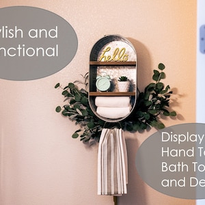 May include: A galvanised metal shelf with two shelves, a wooden top, and a metal sign that says "hello". The shelf is decorated with a green plant, a clock, and towels. The shelf is mounted on a wall with a white towel hanging below it.