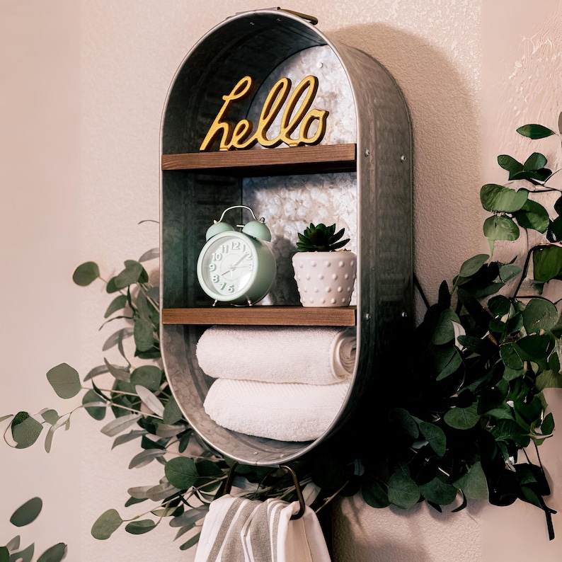 May include: A galvanised metal shelf with two shelves and a hook. The top shelf has the word "hello" in yellow letters. The middle shelf has a green alarm clock and a small potted plant. The bottom shelf has two white towels. The shelf is mounted on a white wall.