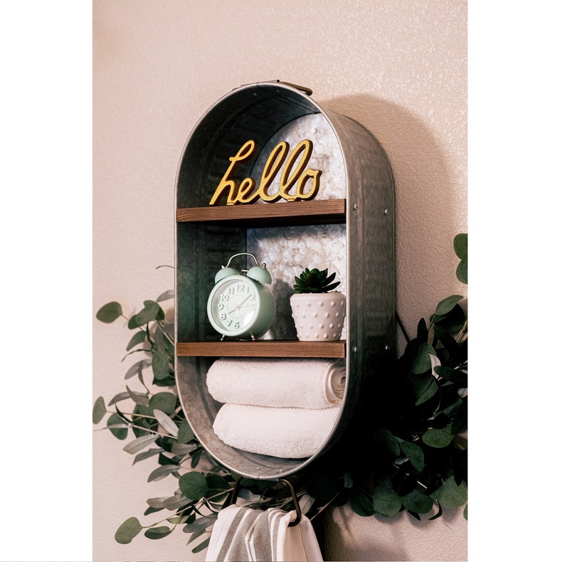 May include: A rustic metal shelf with two shelves and a hook. The shelf is oval shaped and has a galvanised metal finish. The top shelf has a wooden trim and the word "hello" in yellow letters. The bottom shelf has a green alarm clock and a small potted plant. The hook is holding a white and grey striped towel.
