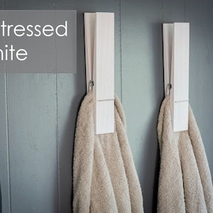 Puede incluir: Dos pinzas de ropa de madera blanca montadas en una pared gris, cada una sujetando una toalla beige. El texto "Distressed White" es visible en la esquina superior izquierda de la imagen.